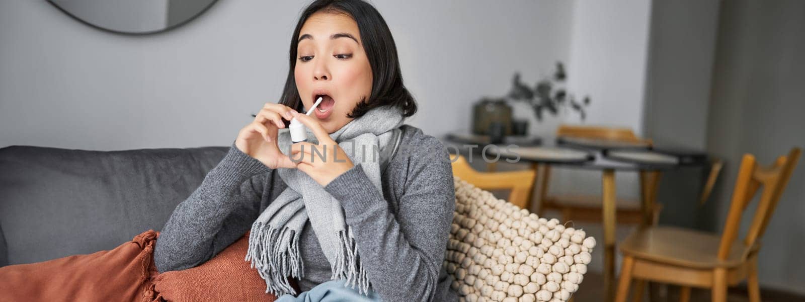 Ill woman using spray from sore throat, treating her cold or flu, staying at home on sick leave, sitting on sofa in living room.