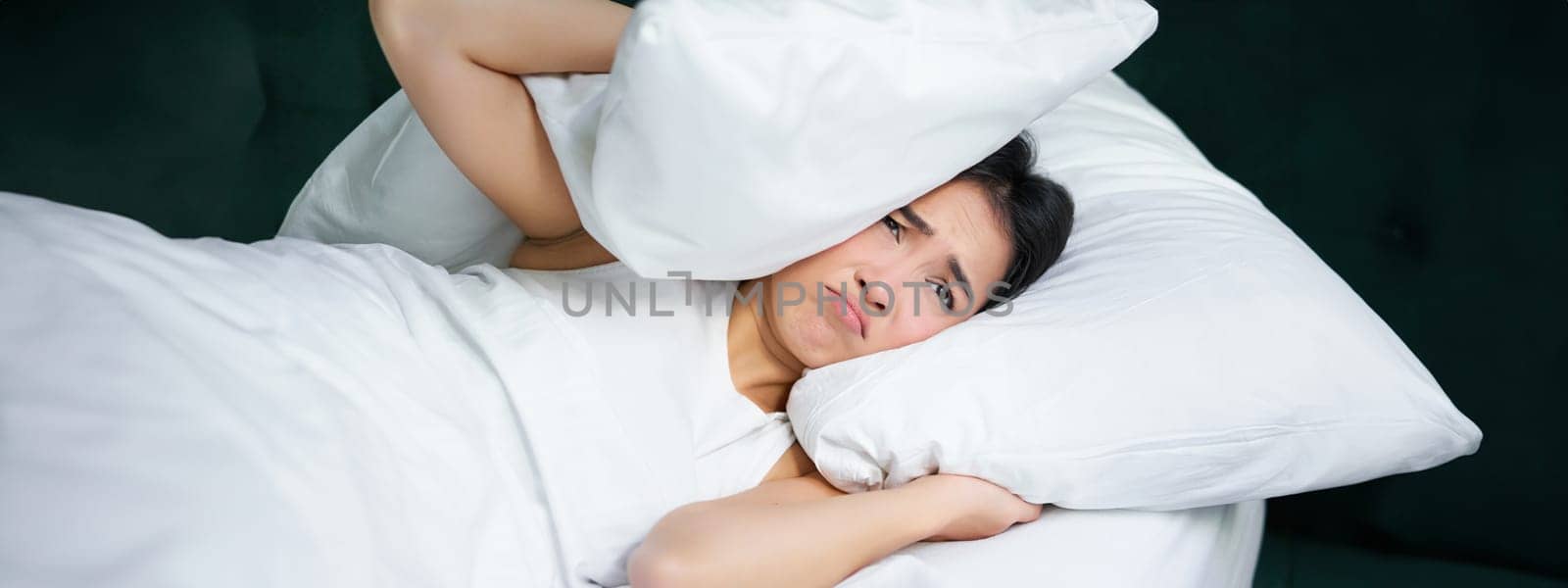 Sad girl with insomnia. Young asian woman lying in bed, cover her ears with pillow, cant sleep, partner snors, loud noise at night disturbing her.