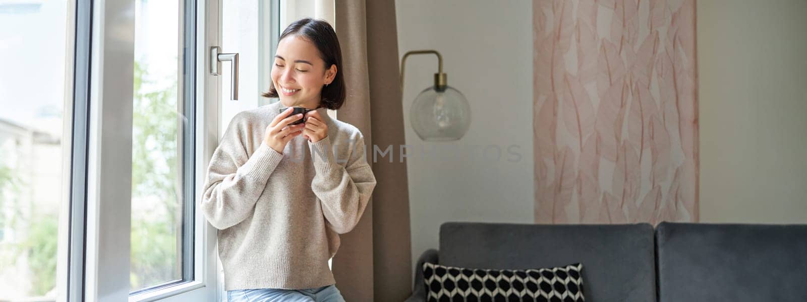 Portrait of young asian woman sitting near window and looking outside, drinking hot coffee from espresso cup and enjoying her cozy day off at home.