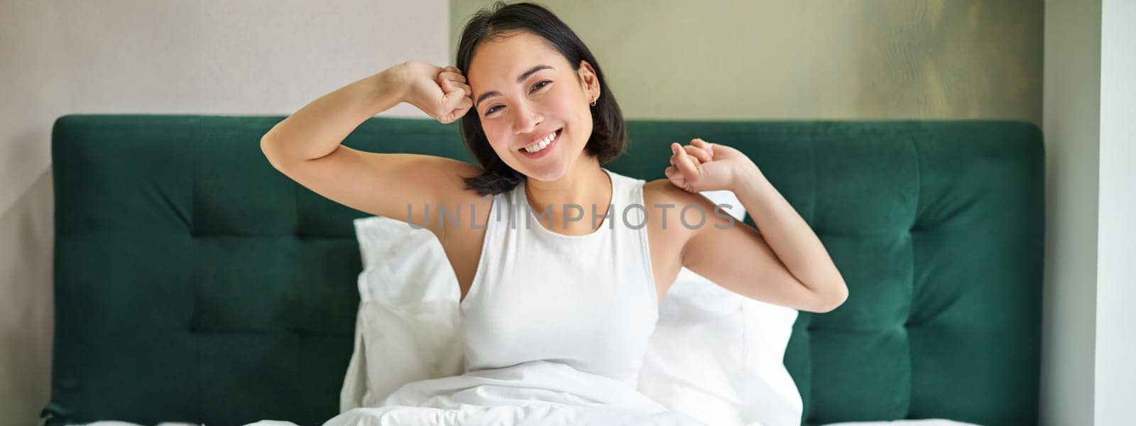 Cute korean girl in white tank top, wakes up in her bedroom, lying in bed in morning, stretching hands and smiles.