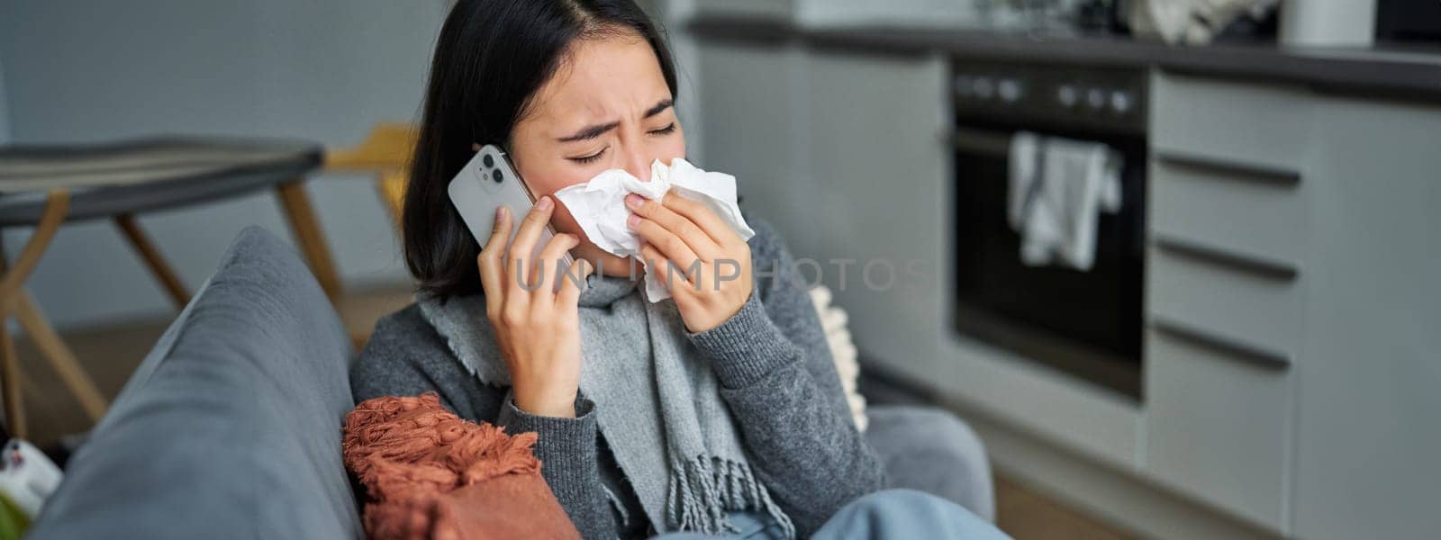 Portrait of ill young korean woman feeling sick, sneezing and holding napkin, staying at home ill, caught cold. Talking on mobile phone by Benzoix