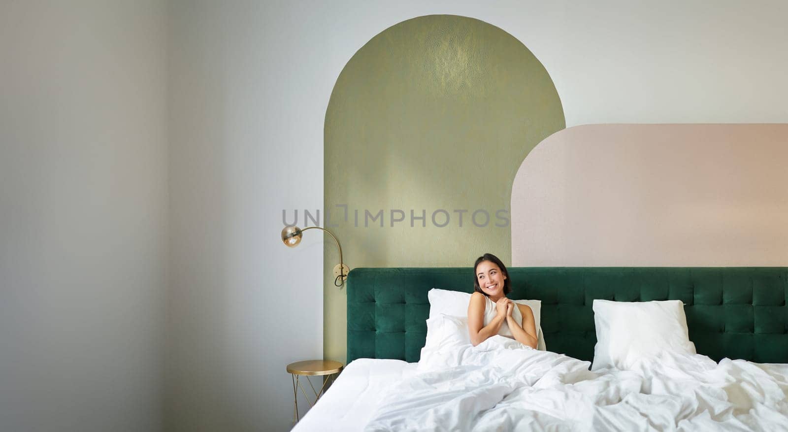 Vertical shot of beautiful asian girl lying in bedroom with sophisticated interrior, smiling and looking happy in morning, spending time in bed by Benzoix