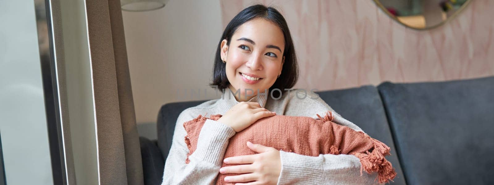 Women and wellbeing concept. Young woman in warm clothes, sits at home, hugs pillow on sofa and smiles pleased, thinks of smth.