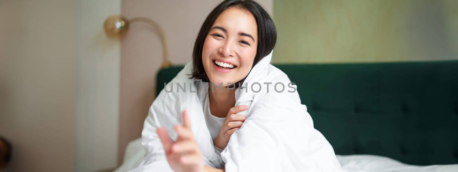 Happy beautiful asian woman covered with blanket, sits on bed in warm duvet, tries to reach camera with a hand, smiling playfully. Hotel bedroom, comfort and apartments concept.