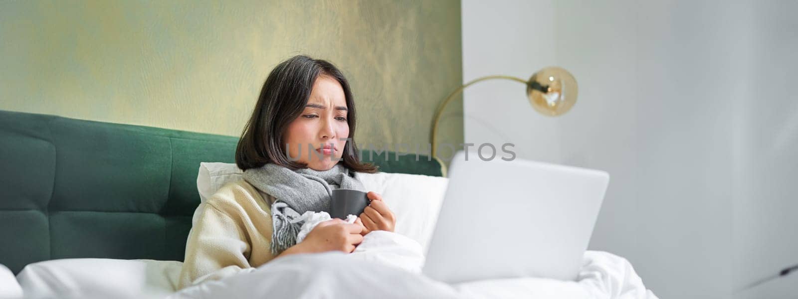 Portrait of young asian woman lying sick in bed, staying at home with cold or flu, watching videos, using laptop in her bedroom by Benzoix