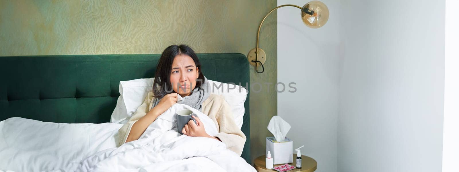 Covid-19, flu and vaccination concept. Korean girl lying in bed with cold, catching influenza, drinking hot tea and using prescribed medication by Benzoix