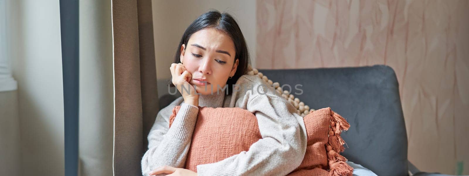 Women and wellbeing concept. Portrait of sad and gloomy asian woman thinking of something unhappy, hugging pillow on sofa and expressing distress by Benzoix