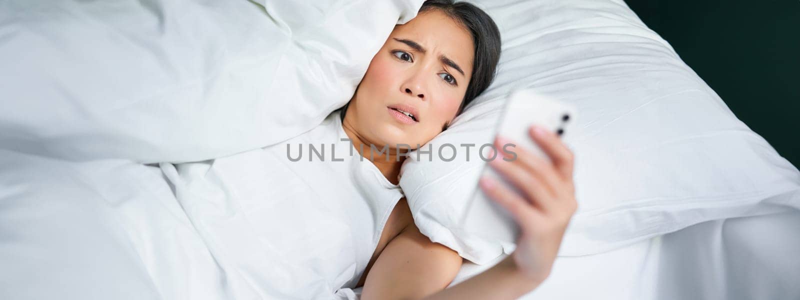 Portrait of asian woman waking up in bed, looking shocked at mobile phone, realise she overslept by Benzoix