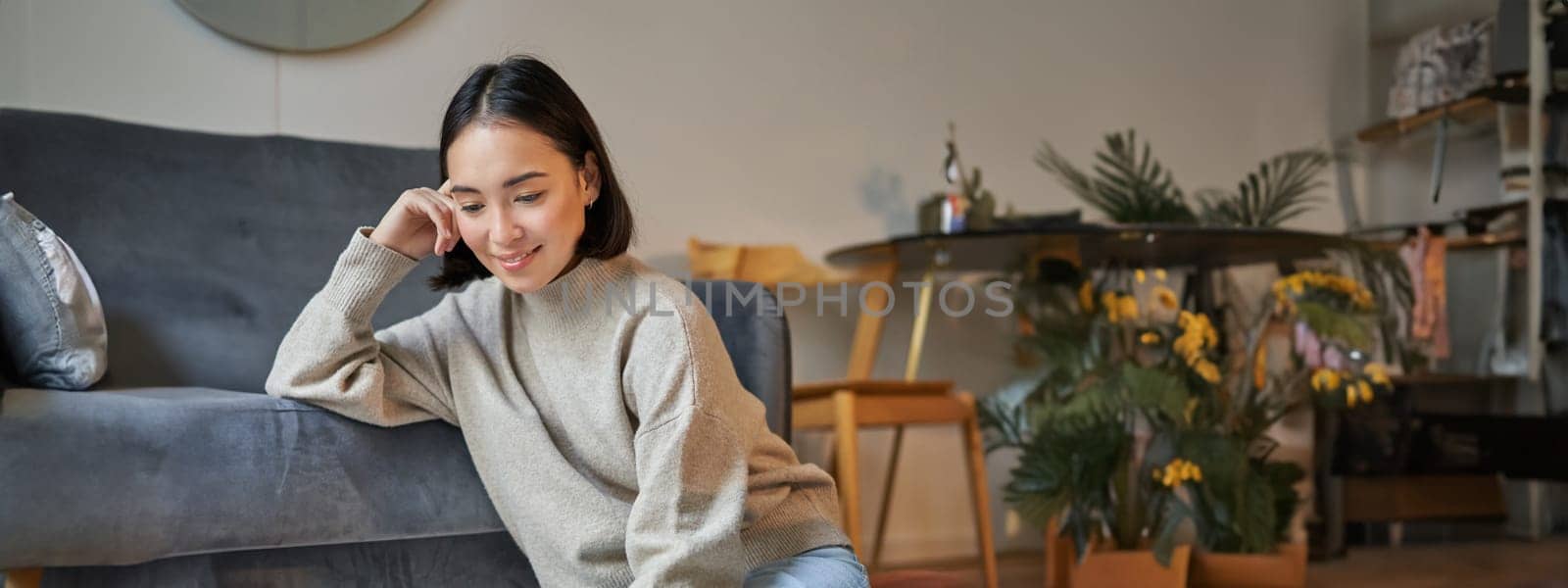 Vertical shot of young woman in cozy home working on laptop, using smartphone and drinking coffee, sitting on floor near sofa.