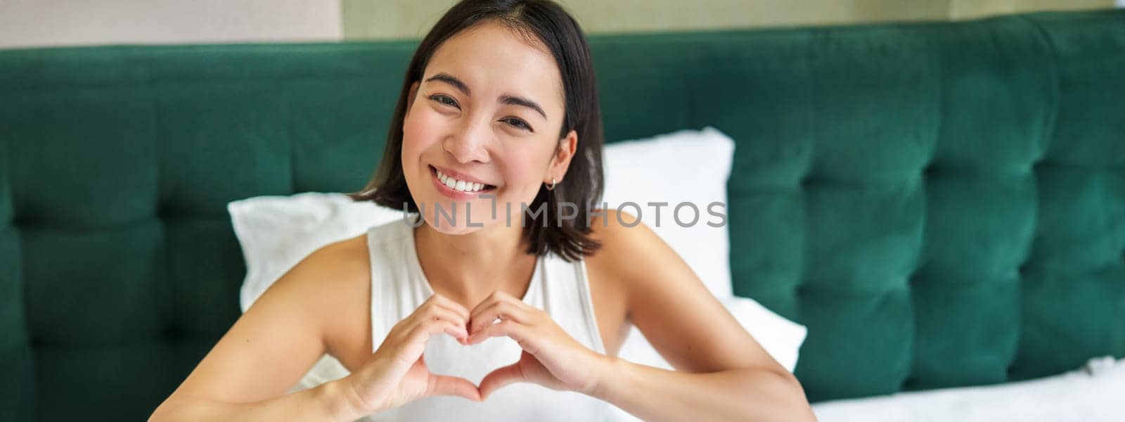 Cute asian girl shows heart, love sign, lying in bed and smiling, enjoying mornings, lazy days in bedroom by Benzoix