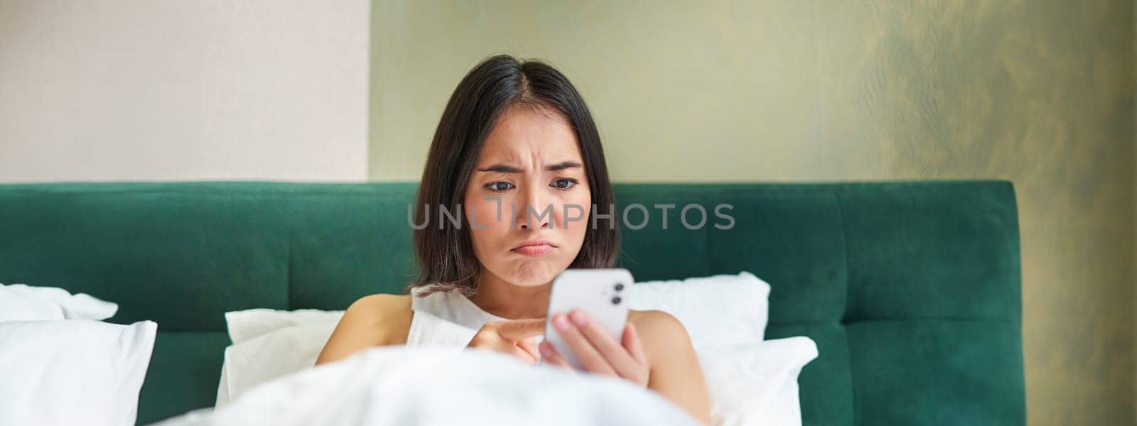 Close up portrait of grumpy asian girl lying in bed, grimacing and pouting from complicated issue on her mobile phone, reading smartphone app by Benzoix