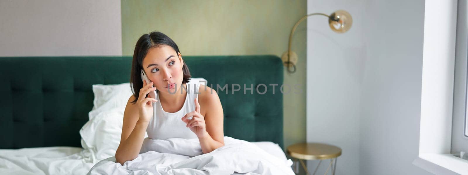 Beautiful young korean woman calling, having a phone call in bed, talking with friend while staying in bedroom.