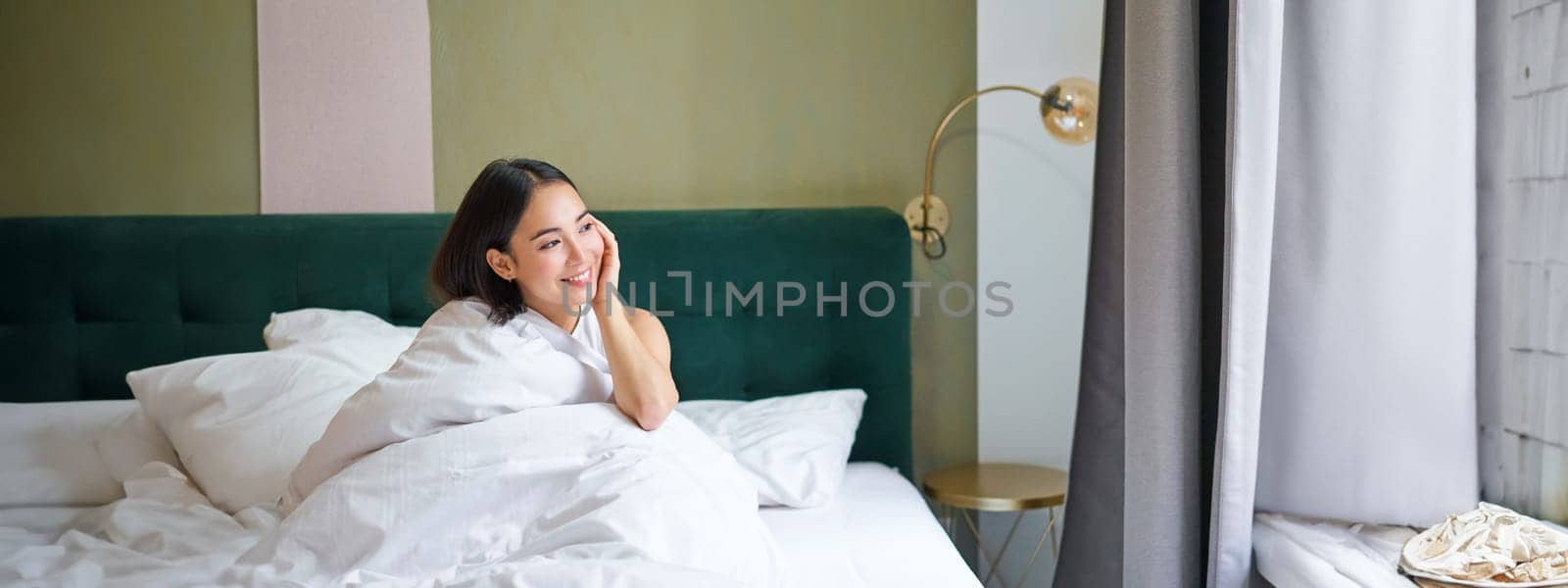 Beautiful sleepy asian girl wakes up in her bed with cozy white blankets, looking outside window with pleased smile. Lifestyle concept by Benzoix