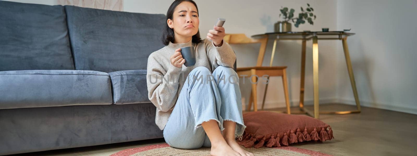 Portrait of girl watching television at home, sits on floor near sofa, holds remote and changes channels.