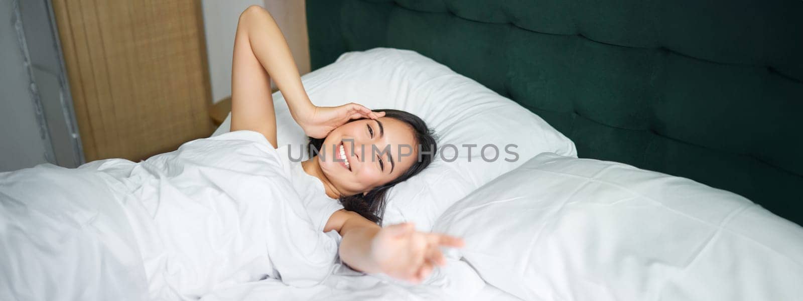 Romantic asian girl wakes up and stretches, smiling happily, reaching her hand, stretching arm to you, lying in bed in white sheets by Benzoix