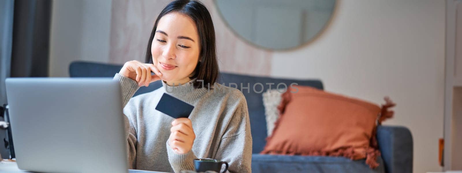 Happy smiling girl with credit card, paying her bills online on computer, doing shopping on her laptop, sitting at home.