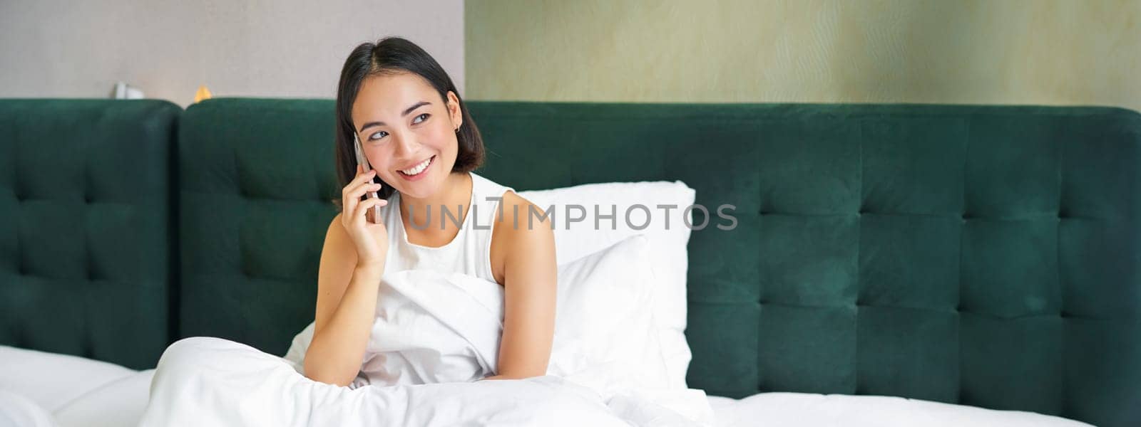 Woman talks on mobile phone in bed. Smiling girl having telephone conversation while relaxing in bedroom by Benzoix