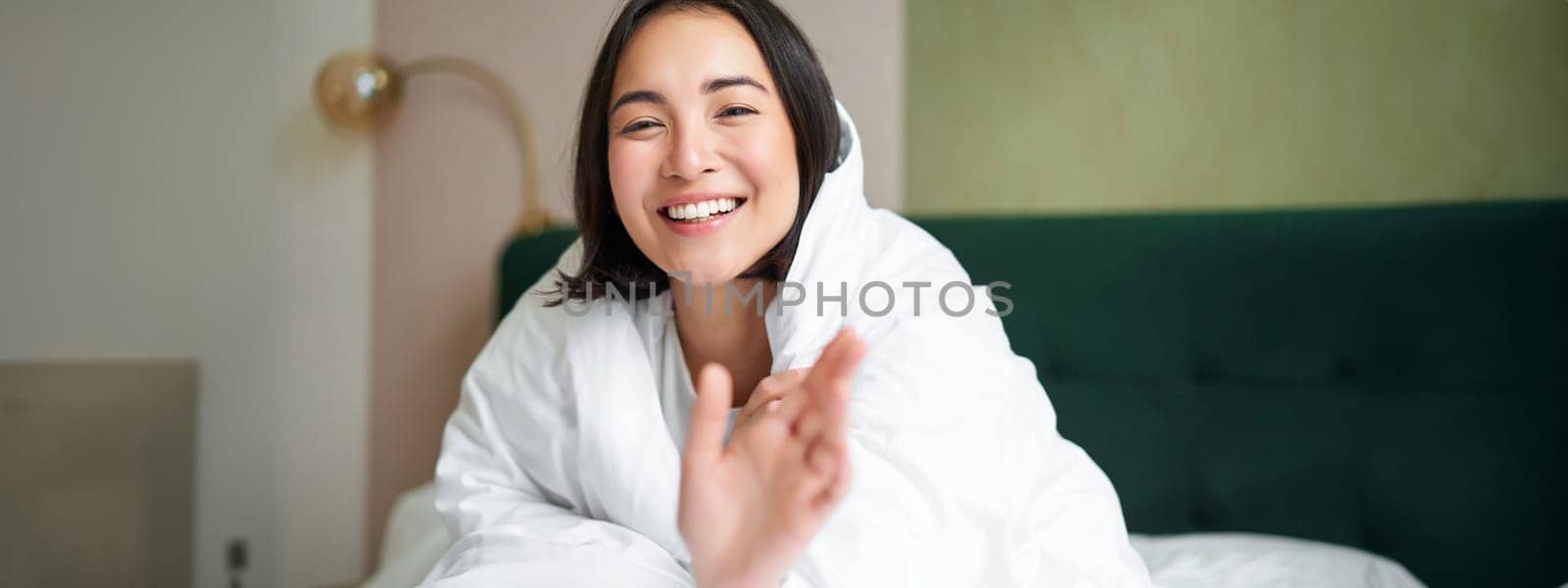 Happy beautiful asian woman covered with blanket, sits on bed in warm duvet, tries to reach camera with a hand, smiling playfully. Hotel bedroom, comfort and apartments concept.