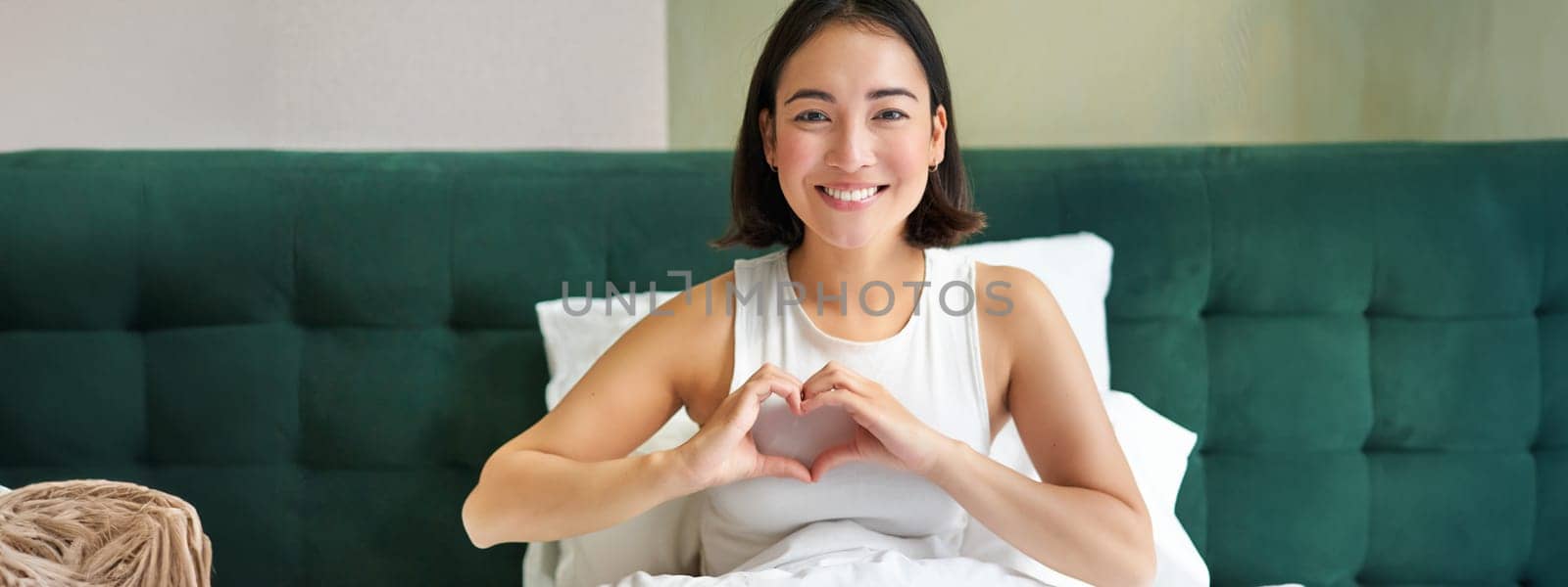 Smiling korean girl shows heart, love sign, spending time in bedroom, lying in bed in morning, covered with white duvets by Benzoix