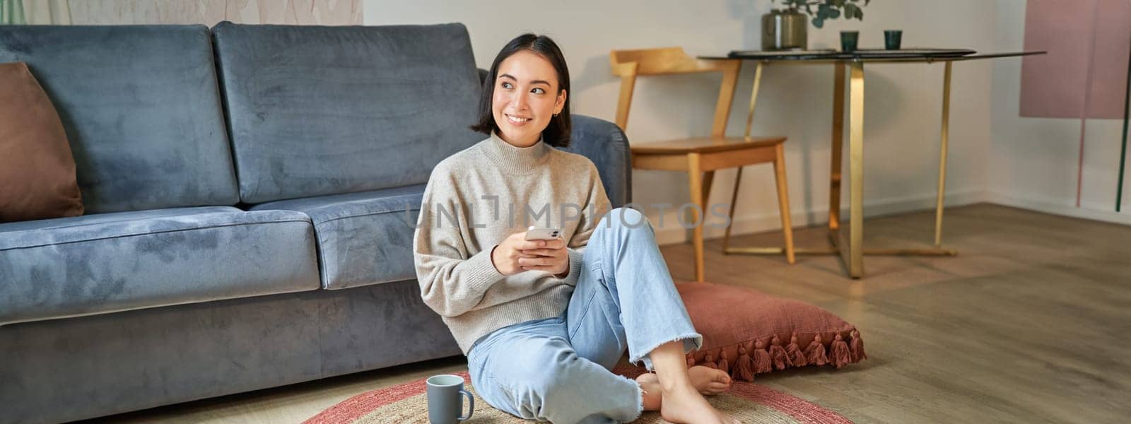 Image of stylish young woman in modern house, using mobile phone, sitting on floor and holding smartphone, drinking from cup.