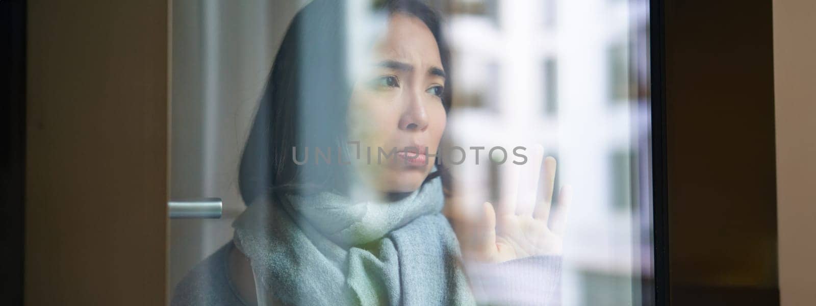 Portrait of sad ill korean woman looking out of the window, staying at home due to coronavirus, being sick and cant leave house.