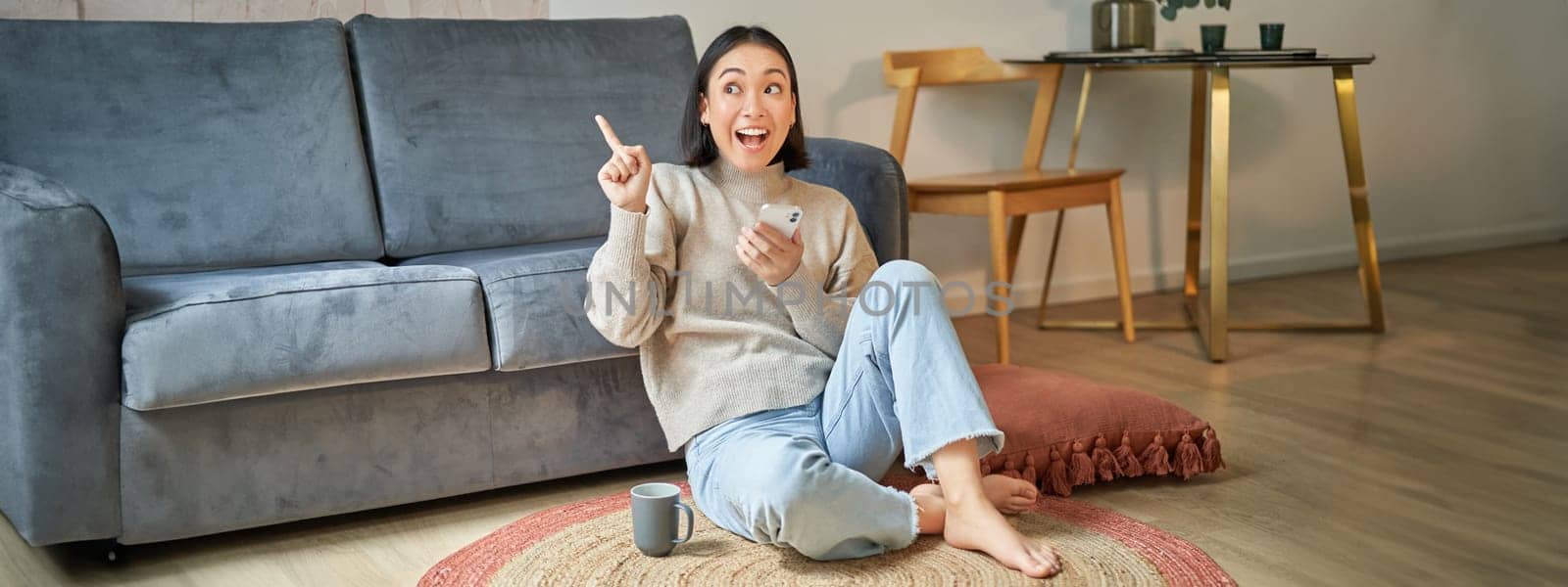 Portrait of modern young korean woman sitting on floor, pointing finger at copy space, showing banner or advertisement, holding smartphone as using app.