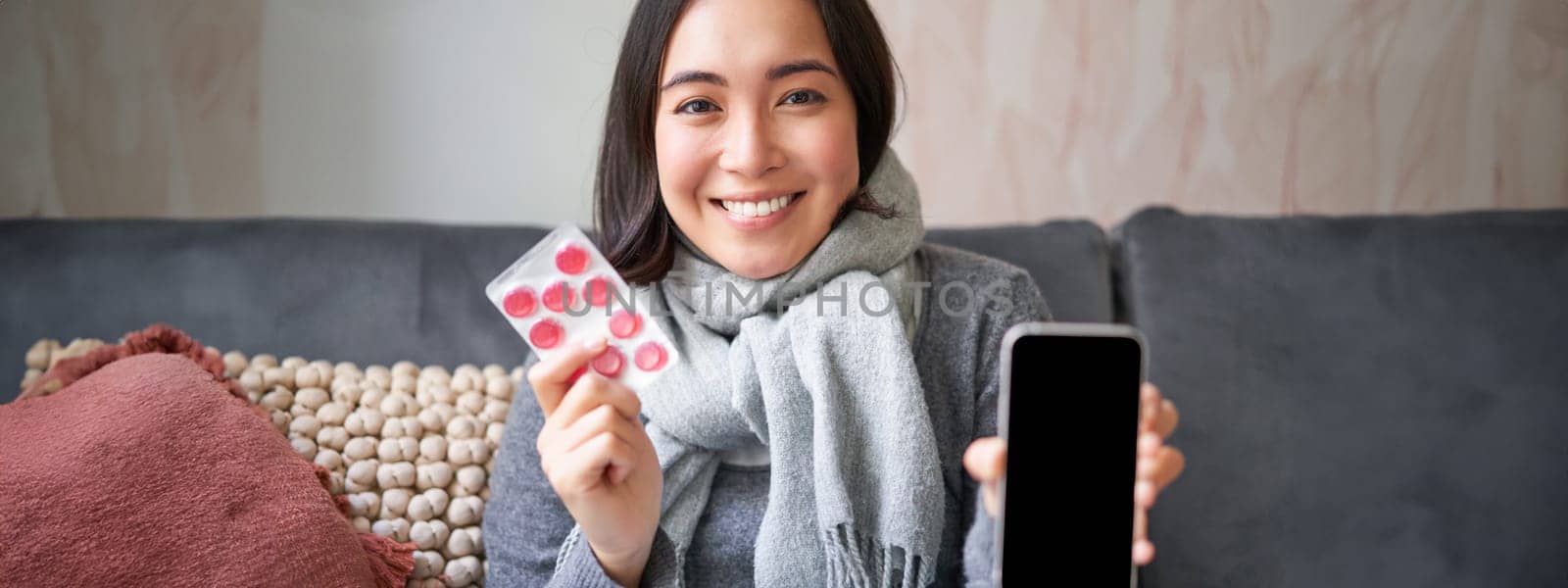 Smiling korean girl shows smartphone screen, medication in hands, feeling sick and staying at home, using online GP doctor app while on sick leave by Benzoix