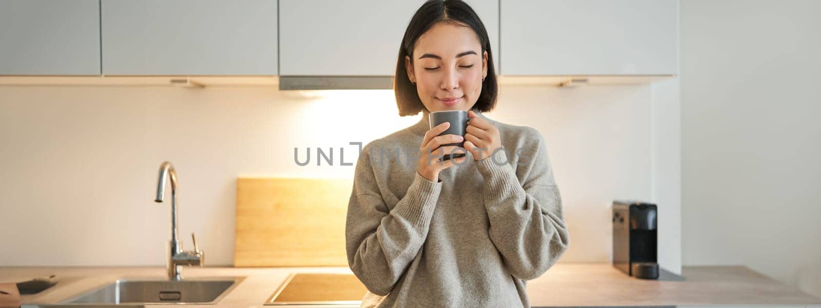 Portrait of smiling asian woman standing on her kitchen, drinking coffee and looking at camera, concept of cozy and comfortable home by Benzoix