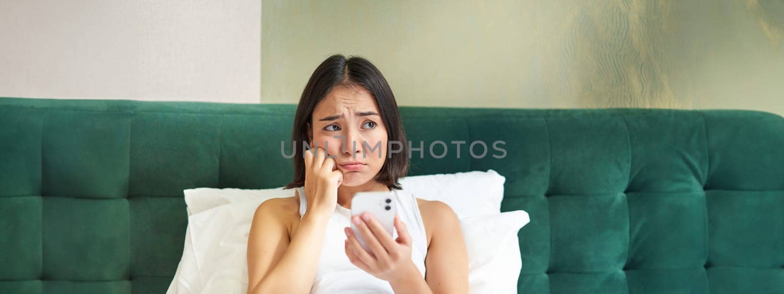 Portrait of sad frowning korean girl, lying in bed and looking at smartphone with upset, disappointed face expression, using mobile phone, complaining by Benzoix