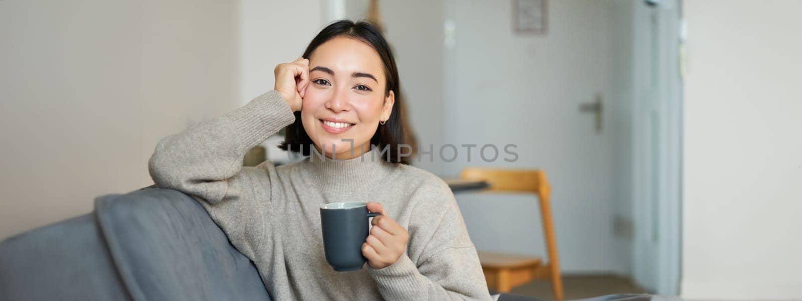 Smiling asian woman sitting at home with cup of coffee, relaxing and feeling warmth, looking outside window, resting on sofa in living room by Benzoix