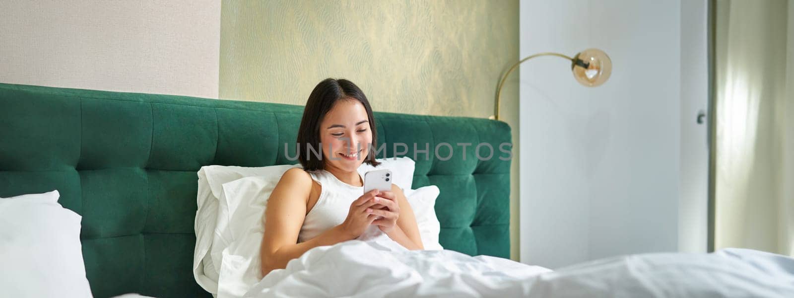 Cute korean girl in bed, holding smartphone, feeling happy and pleased, spending morning in bed, enjoying surfing net on mobile phone.