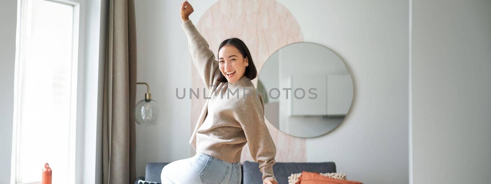 Cute excited asian woman dancing, raising hands up in triumph, celebrating and singing, standing at home in warm jamper by Benzoix