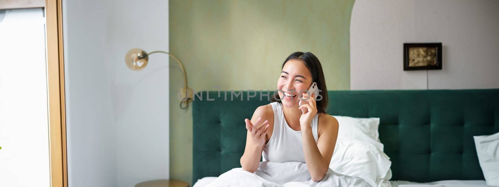 Beautiful smiling asian girl talking on mobile phone, lying in bed with pleased happy face, speaking to someone on telephone.