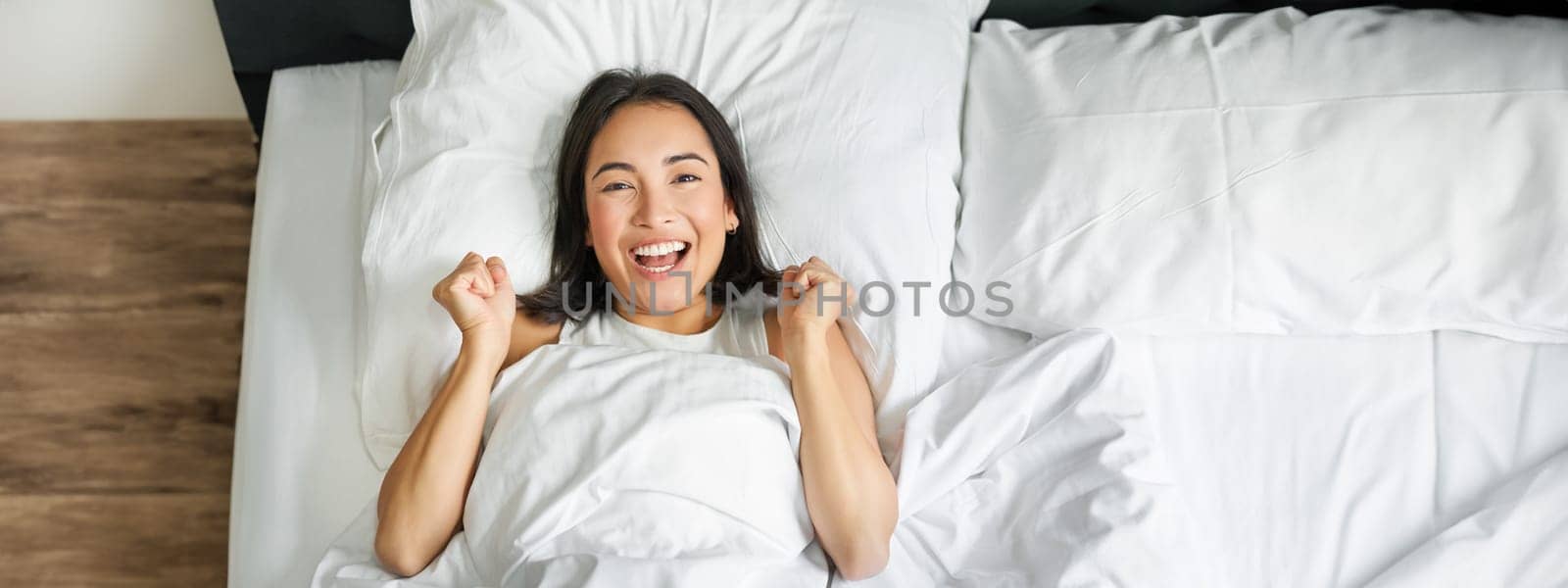 Top view of enthusiastic asian girl cheering, lying in her bed and shouting with joy and excitement, triumphing and smiling, enjoy staying in her bedroom by Benzoix