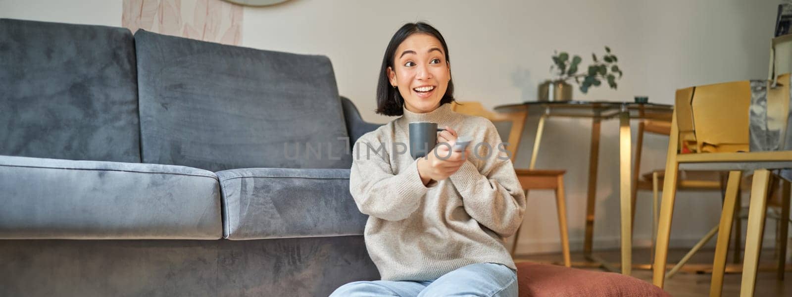 Portrait of smiling young woman resting at home near tv, watching television sitting on floor and drinking coffee from cup.