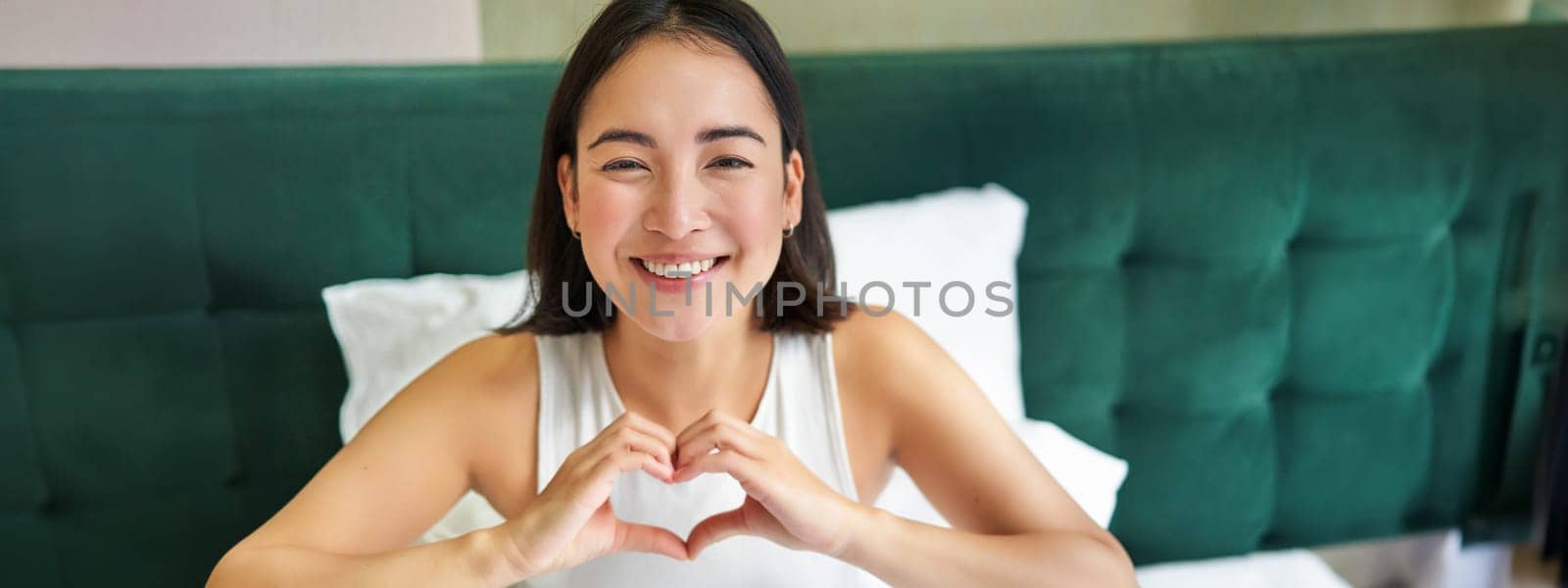 Cute asian girl shows heart, love sign, lying in bed and smiling, enjoying mornings, lazy days in bedroom by Benzoix