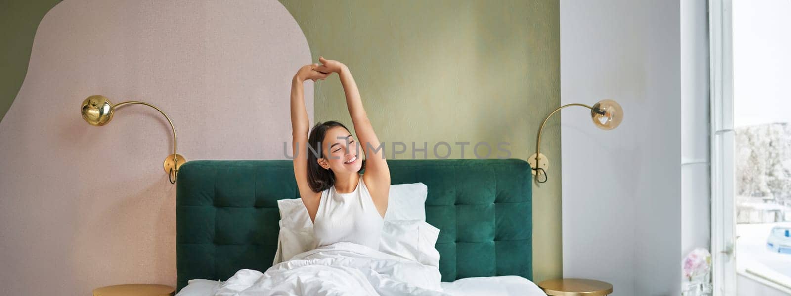 Beautiful asian woman lying in her bedroom on bed, stretching hands and looking outside, waking up from sleep.