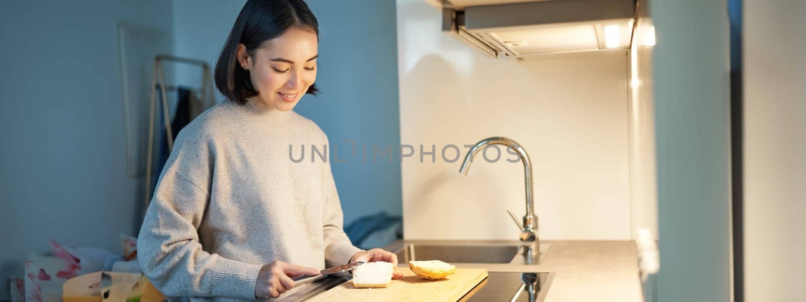 Cute asian woman making herself toast, cut loaf of bread, preparing sandwitch on kitchen by Benzoix