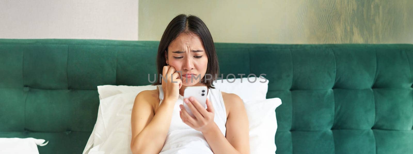 Portrait of sad frowning korean girl, lying in bed and looking at smartphone with upset, disappointed face expression, using mobile phone, complaining by Benzoix