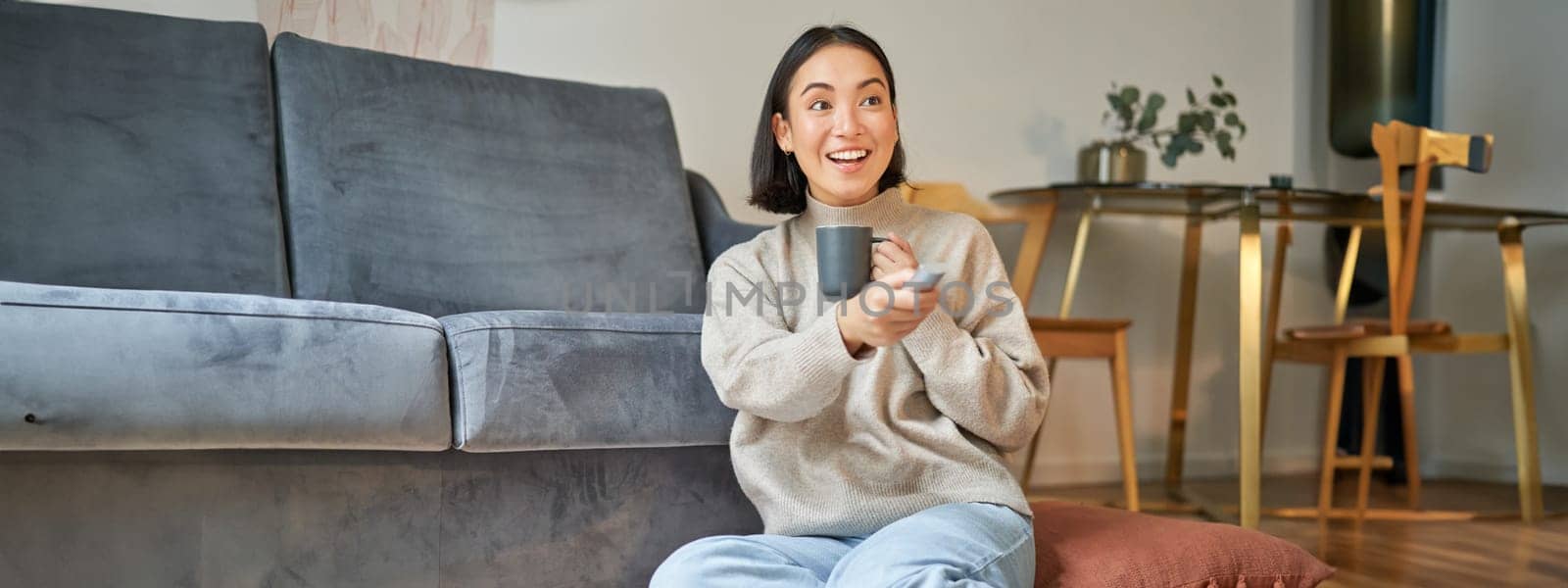 Portrait of smiling young woman resting at home near tv, watching television sitting on floor and drinking coffee from cup.