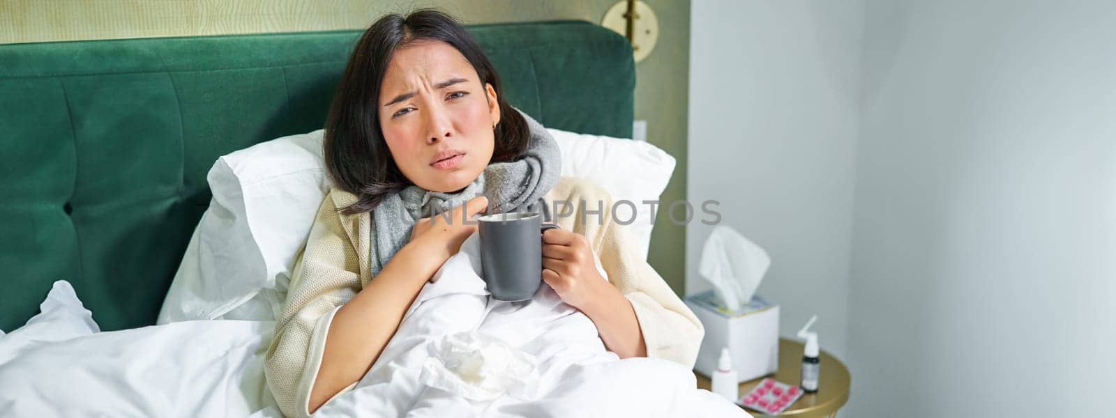 Portrait of couching asian girl in bed, feeling sick, catching cold and staying at home, looking unhealthy, drinking hot tea by Benzoix