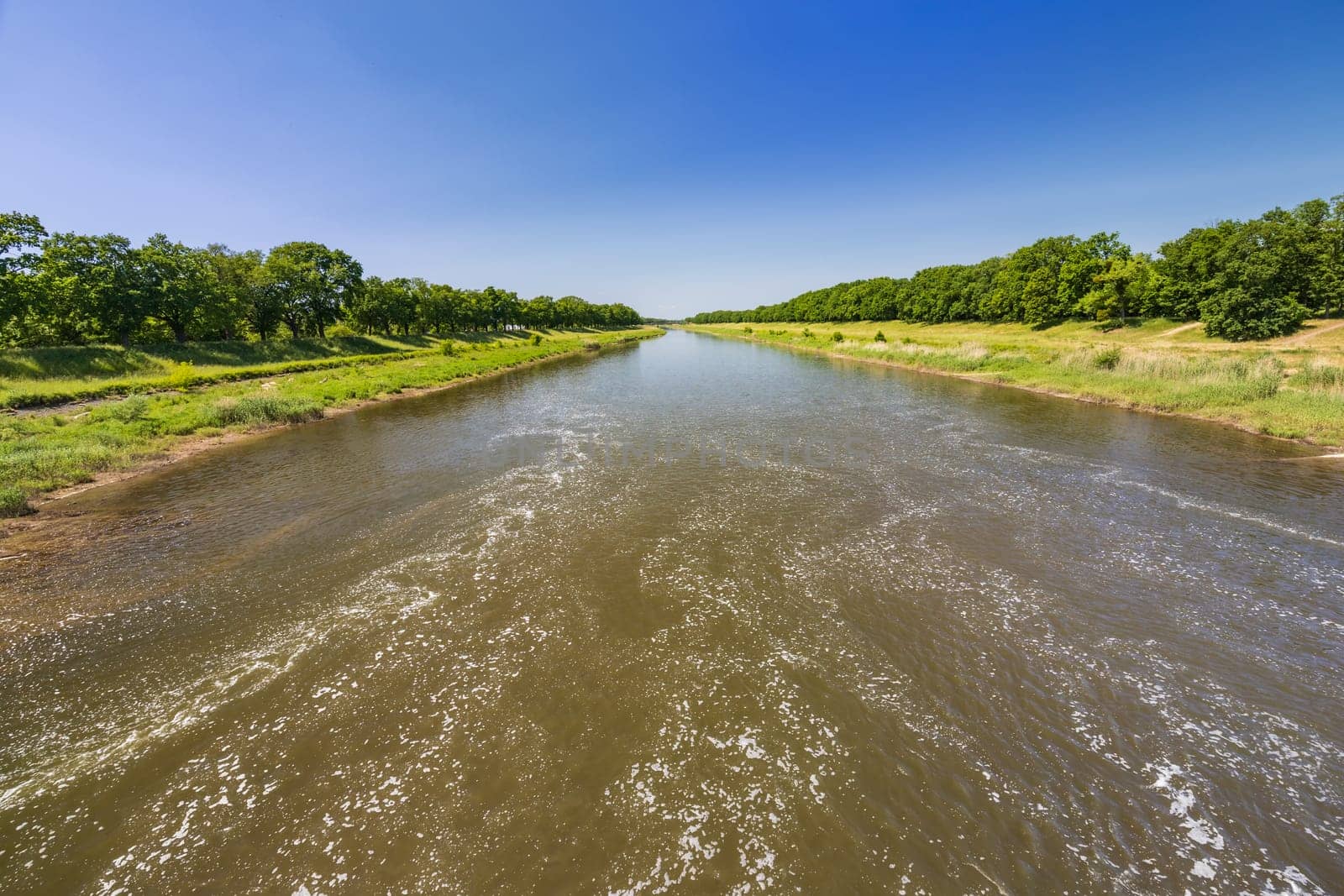 Beautiful landscape of long dirty river with green trees and bushes on both sides seen from bridge over water dam and water level by Wierzchu