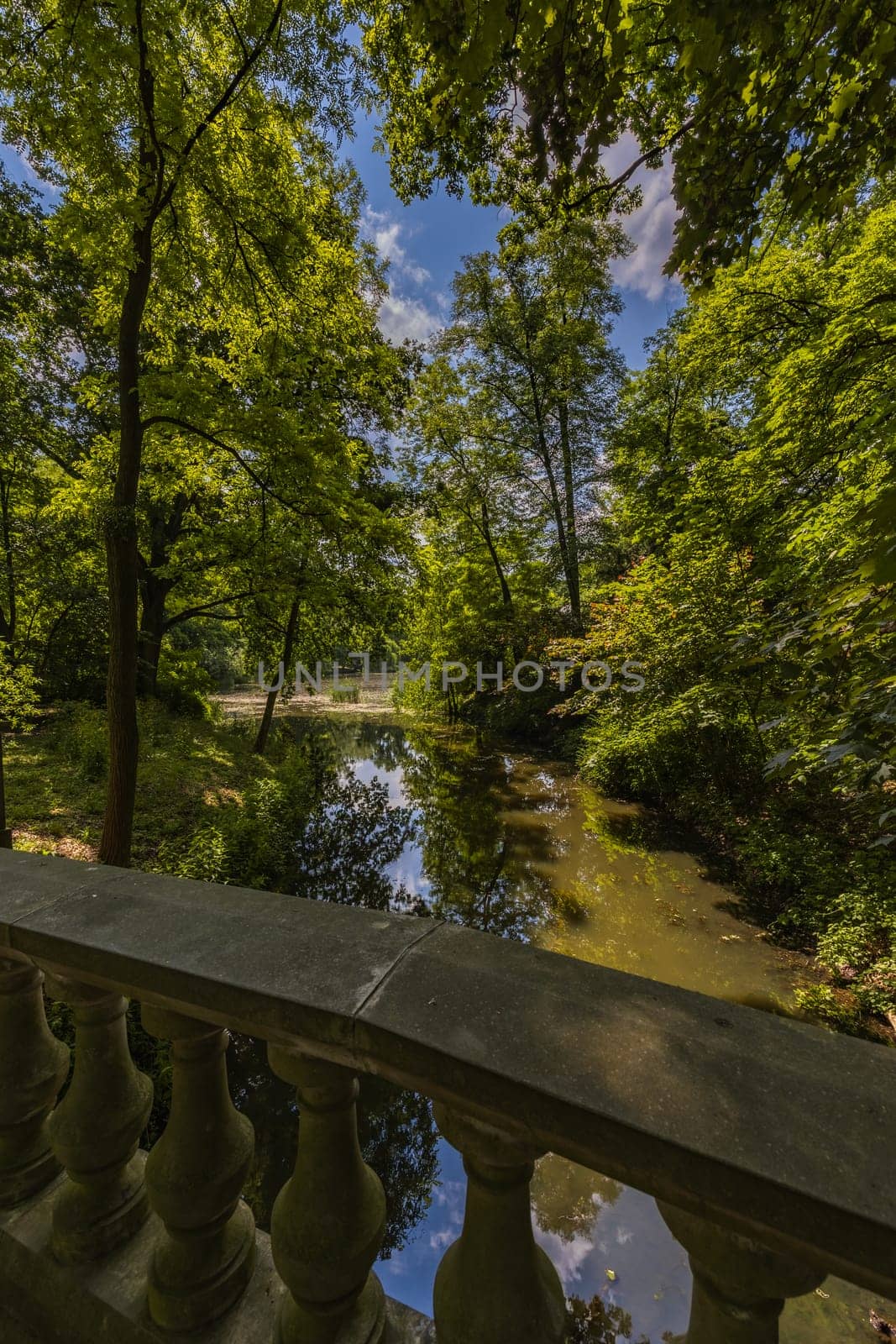 Small stream in beautiful park with colorful surroundings seen from small concrete bridge with beautiful decorations by Wierzchu