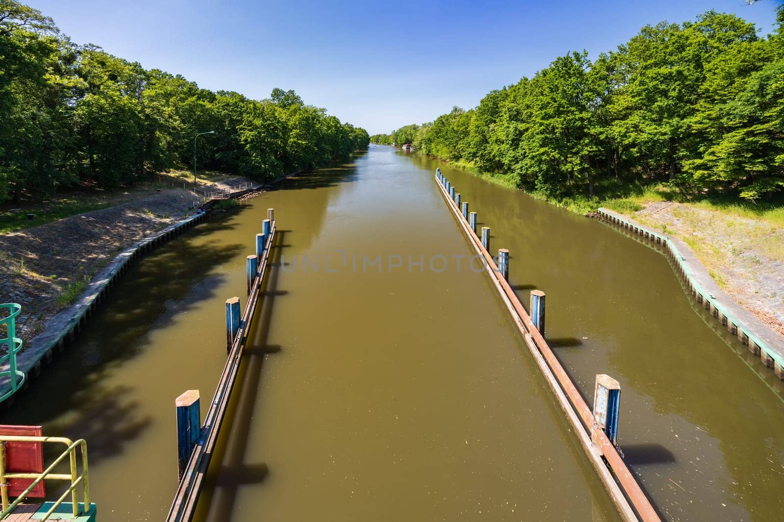 Beautiful view of boat entrance to water level mechanism with dirty water with beautiful green trees and bushes on both sides of river by Wierzchu