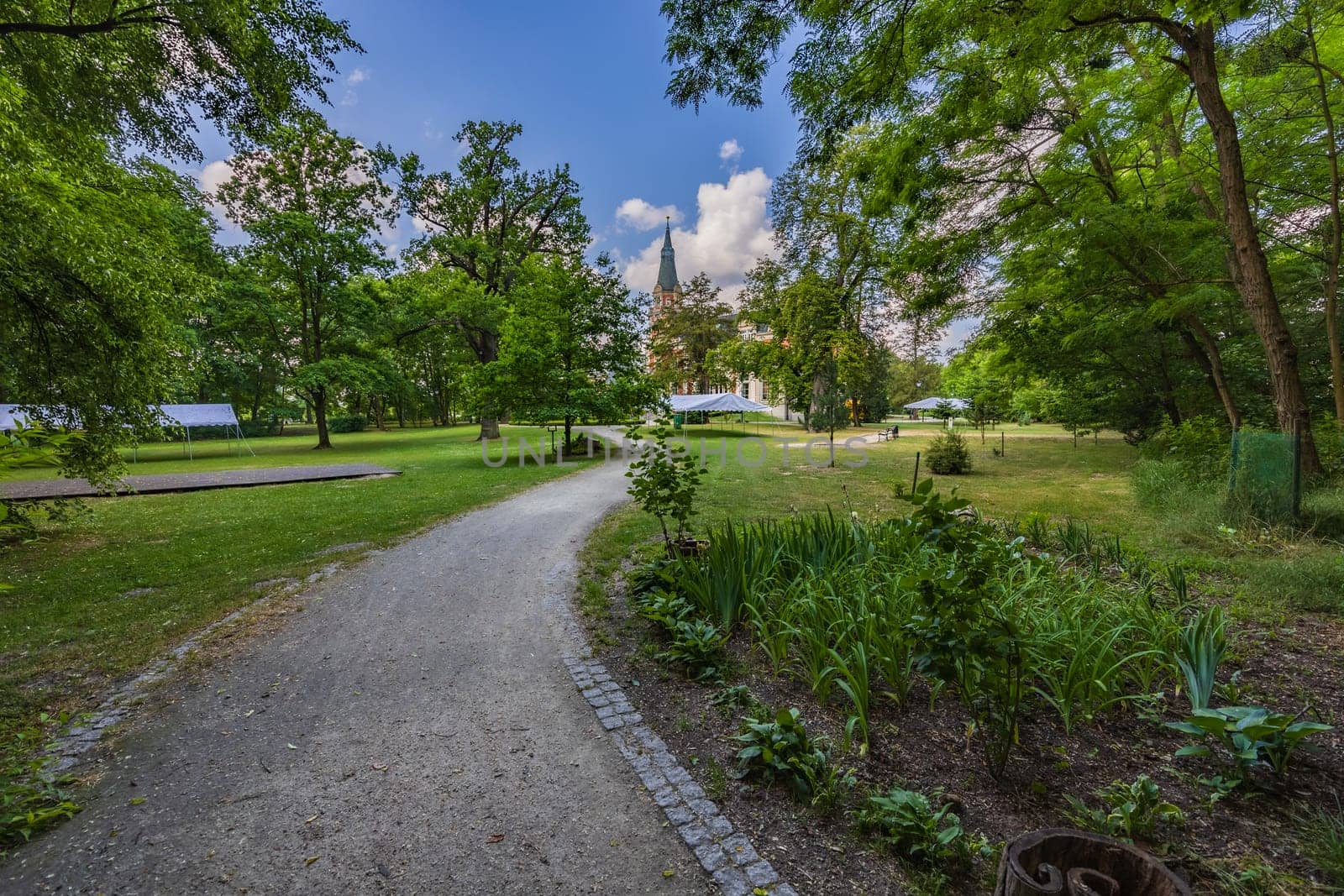 Long curvy path in beautiful green park with tower of building at sunny morning with blue sky and few white clouds by Wierzchu