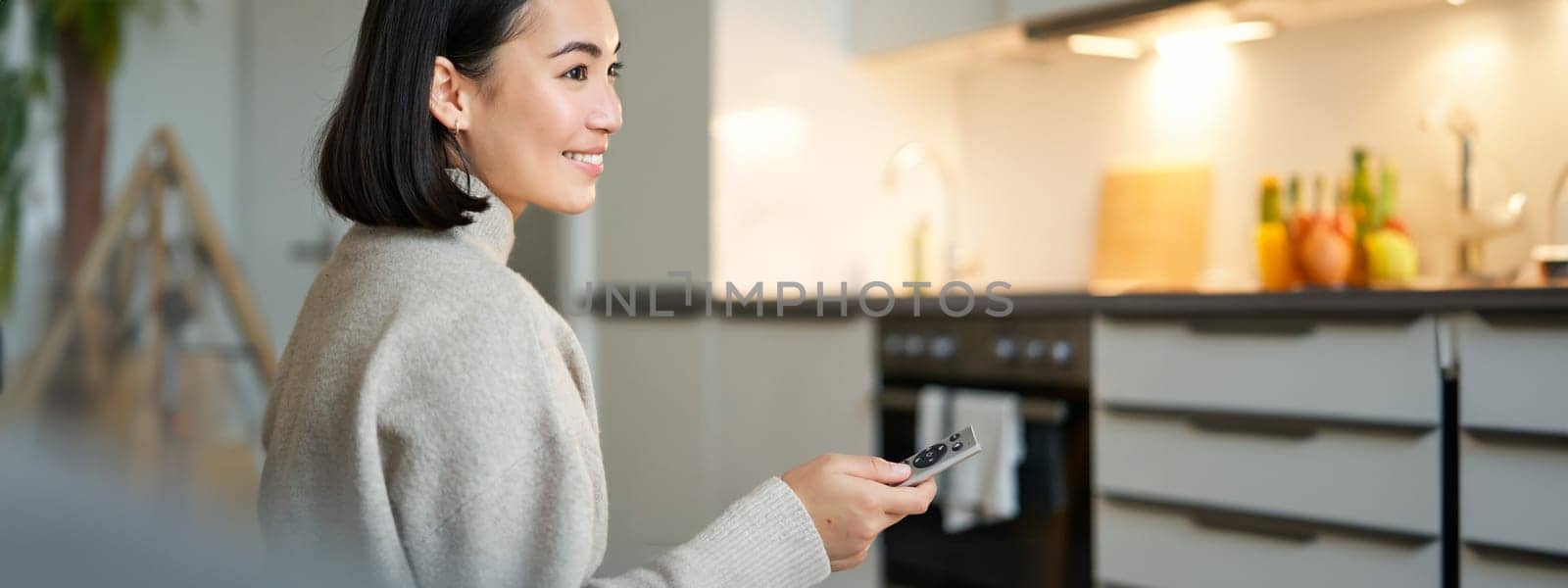 Close up portrait of smiling asian woman watching television, sitting on sofa with remote and switching chanel, looking relaxed by Benzoix