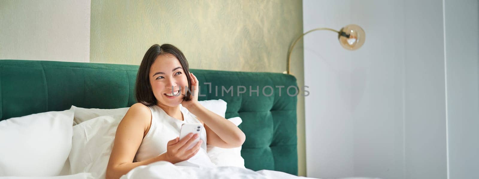 Cute korean girl in bed, holding smartphone, feeling happy and pleased, spending morning in bed, enjoying surfing net on mobile phone.