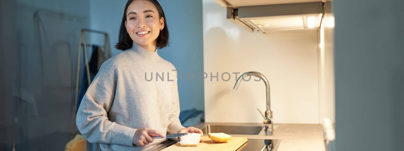 Cute asian woman making herself toast, cut loaf of bread, preparing sandwitch on kitchen.