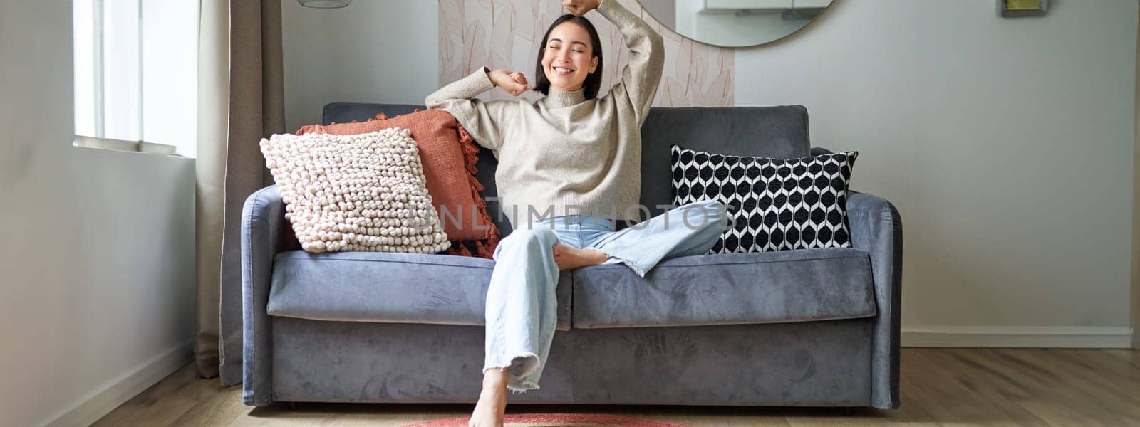 Portrait of happy asian woman feeling lazy, stretching on sofa and smiling pleased, relaxing at home, resting from work.