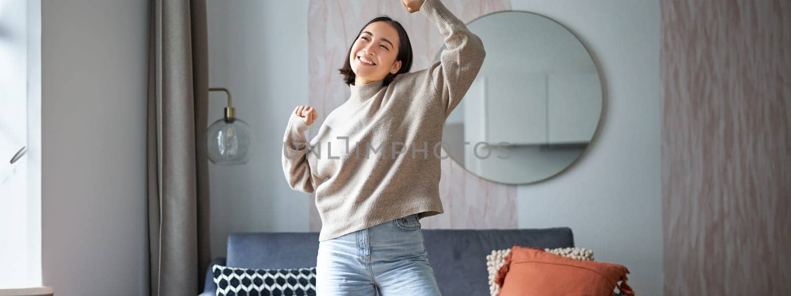 Portrait of happy asian woman dancing, rejoicing and triumphing, feeling upbeat at home by Benzoix